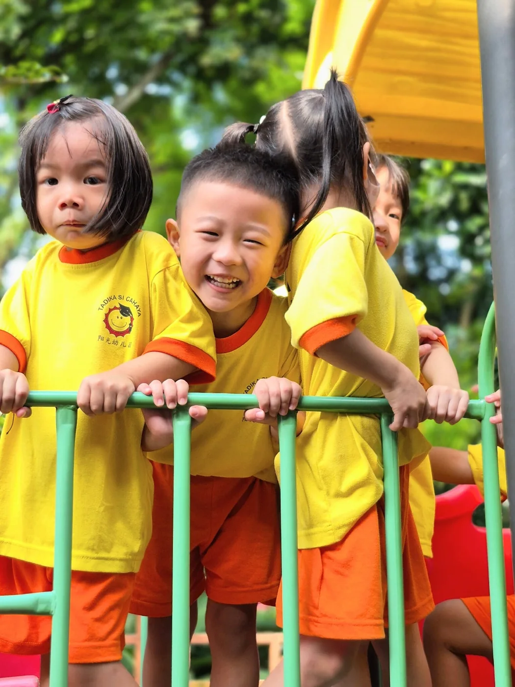 Three children smiling together at an outdoor playground railing.
